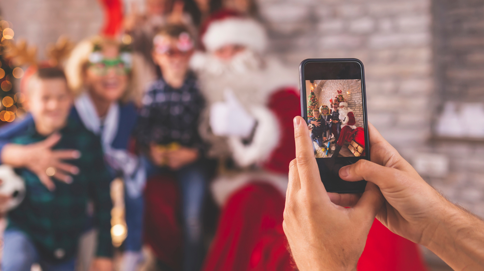 A person holds a smartphone while taking a photo of a family posing with Santa Claus during a holiday celebration.