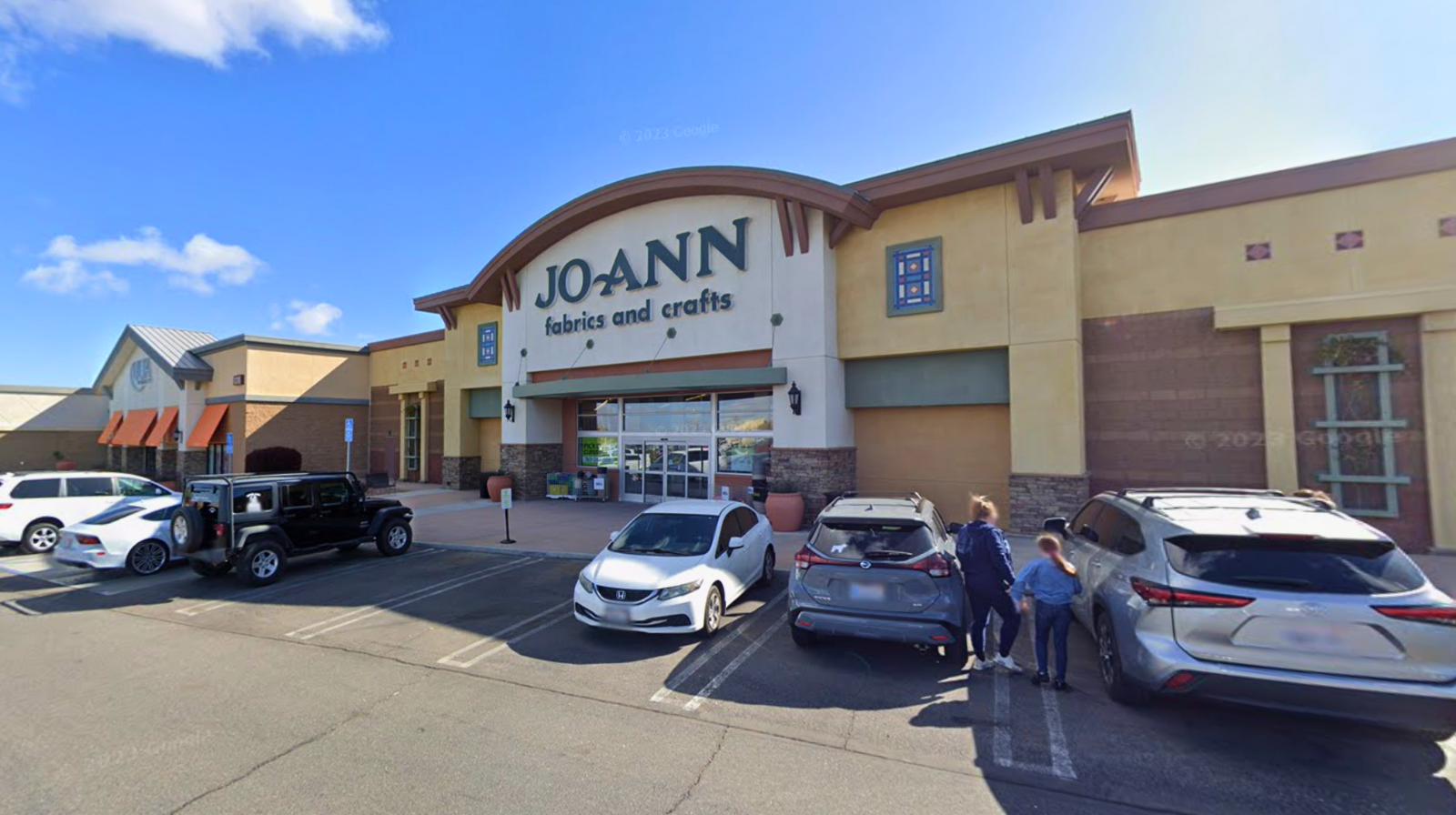 Exterior view of a JOANN Fabrics and Crafts store in a shopping center, showing the storefront sign, parking lot with cars, and customers walking toward the entrance on a sunny day.