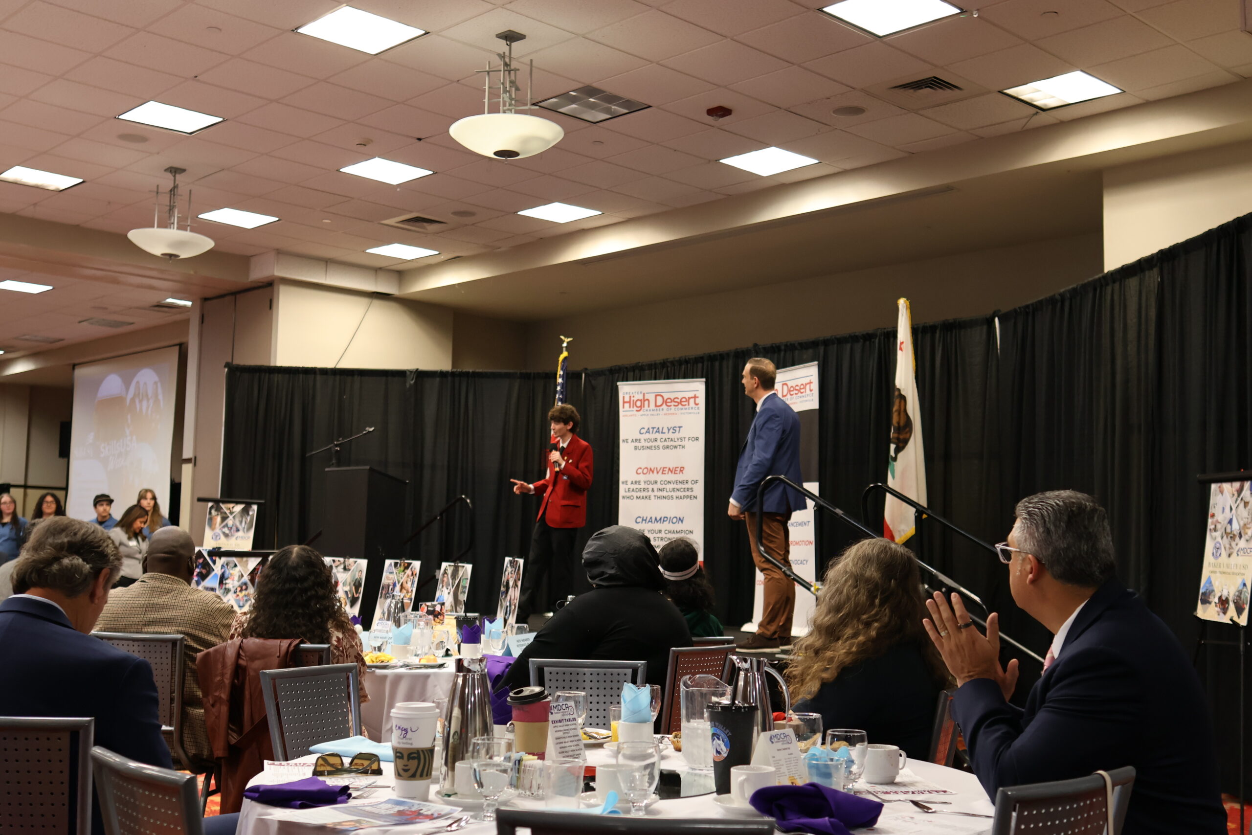 Speakers present on stage at a community business event while attendees sit at round tables and listen, with High Desert Chamber of Commerce signage and flags visible in the background.