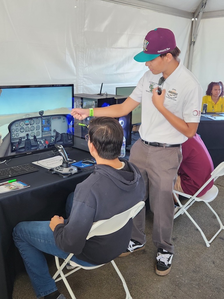 Two aviation students practicing on a flight simulator, one seated at a desk and the other standing and giving instructions.