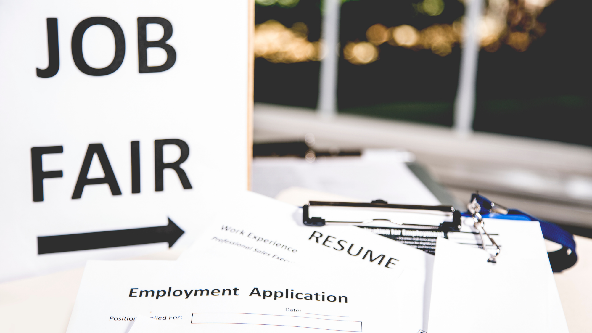 Close-up image of a “Job Fair” sign with an arrow pointing right, alongside a clipboard holding a resume and employment application form.