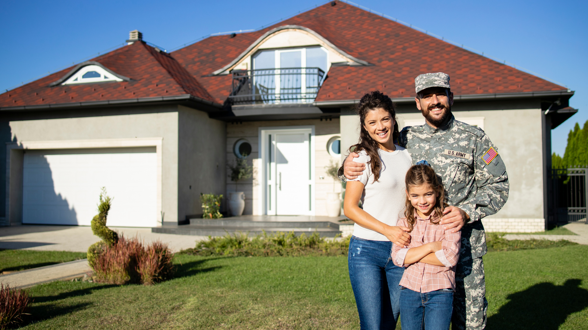 A smiling military service member in uniform stands with his family in front of a suburban home.