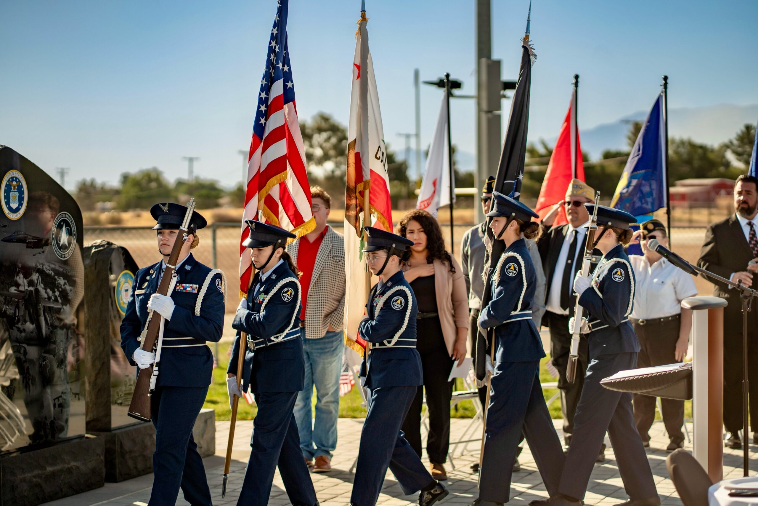 A JROTC color guard in blue dress uniforms marches with American and military flags during an outdoor memorial ceremony, while attendees and veterans stand nearby under a clear sky.