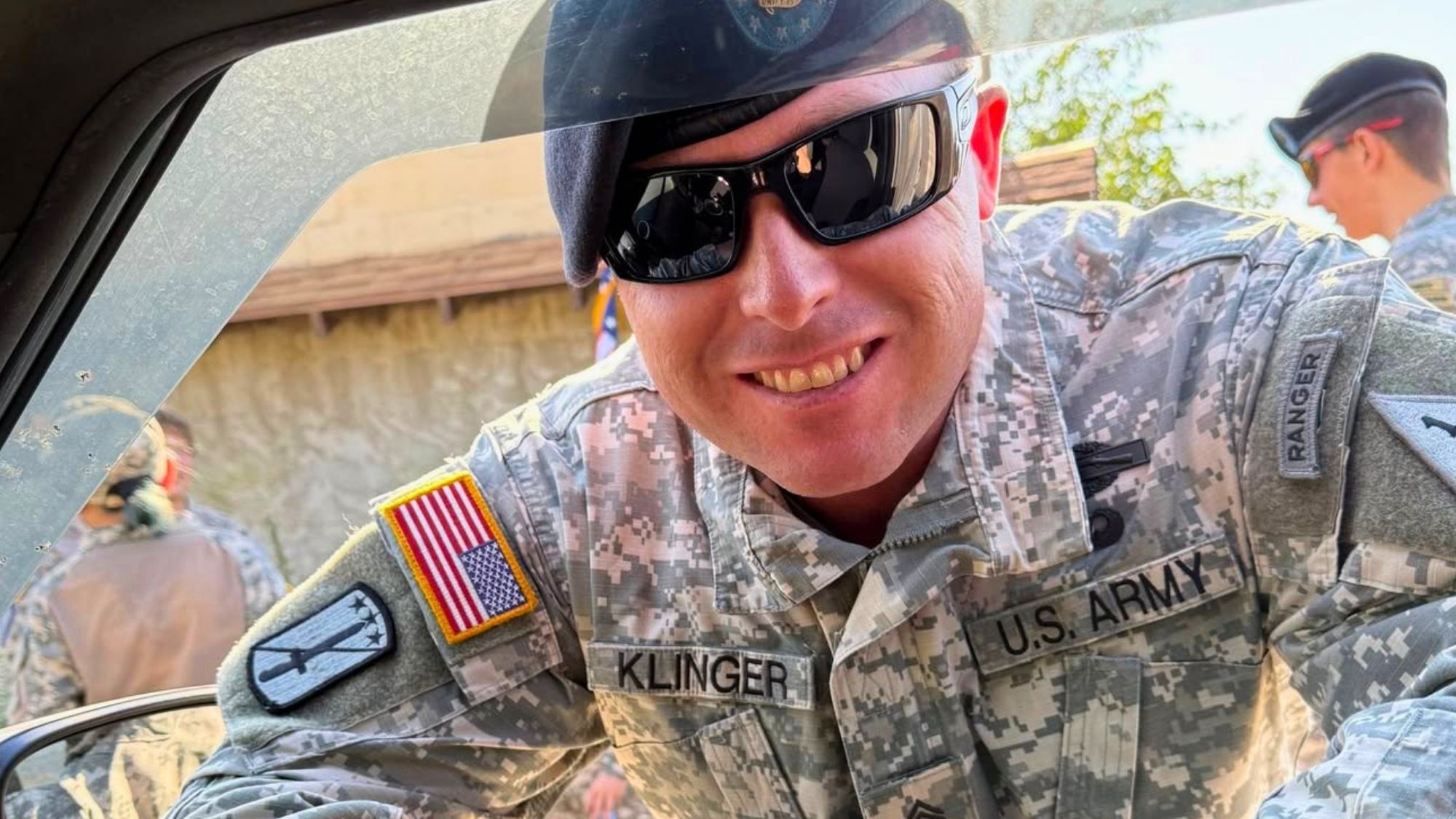 Close-up of a smiling U.S. Army soldier in camouflage uniform and sunglasses leaning into a vehicle window, with other soldiers visible in the background outdoors.