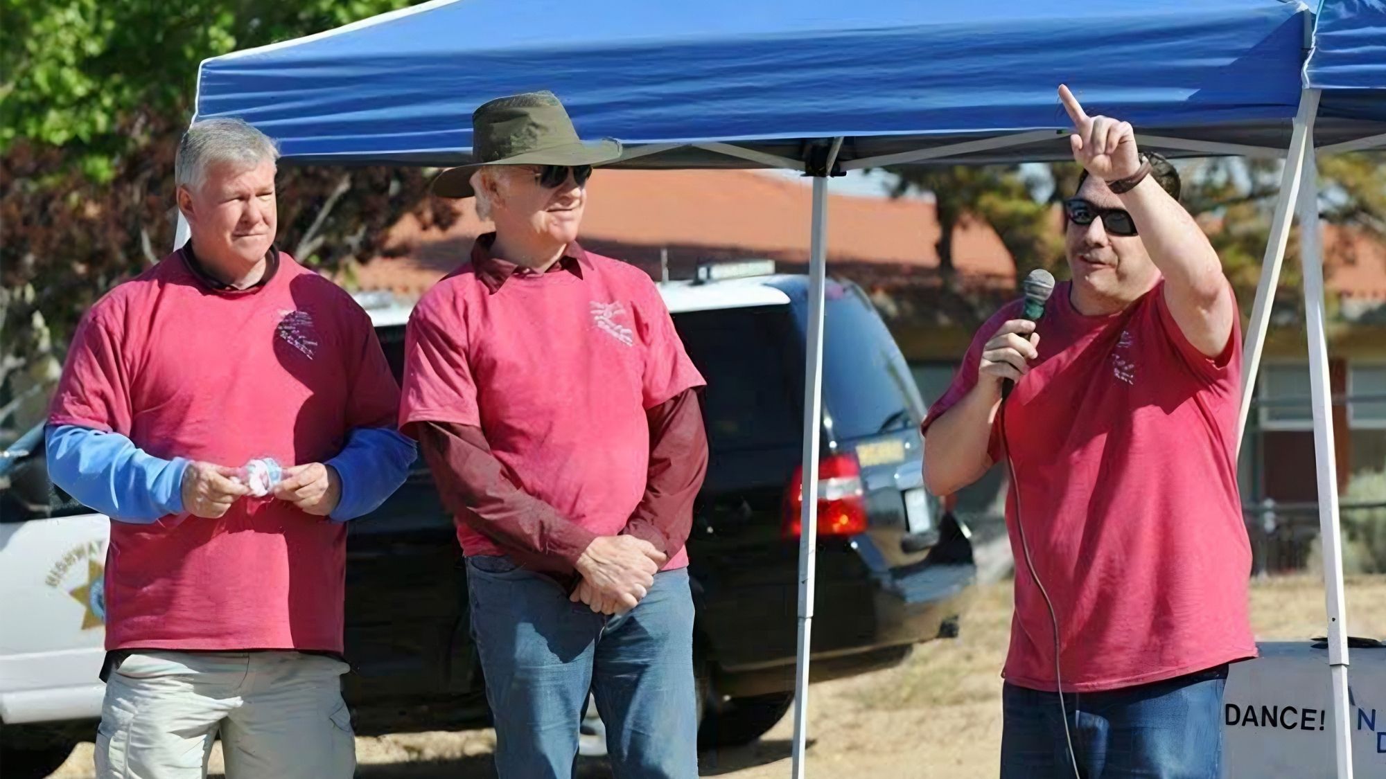 Three men in matching red shirts stand under a canopy at an outdoor event, one speaking into a microphone while gesturing as the others look on.