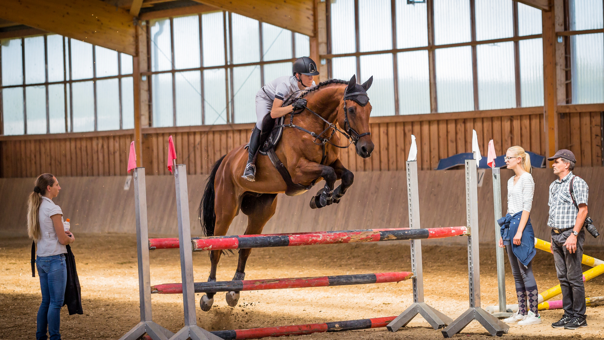 MojaveNarrowsEquestrian Equestrian rider in helmet jumps a horse over a hurdle inside an indoor arena while three people watch from the side.