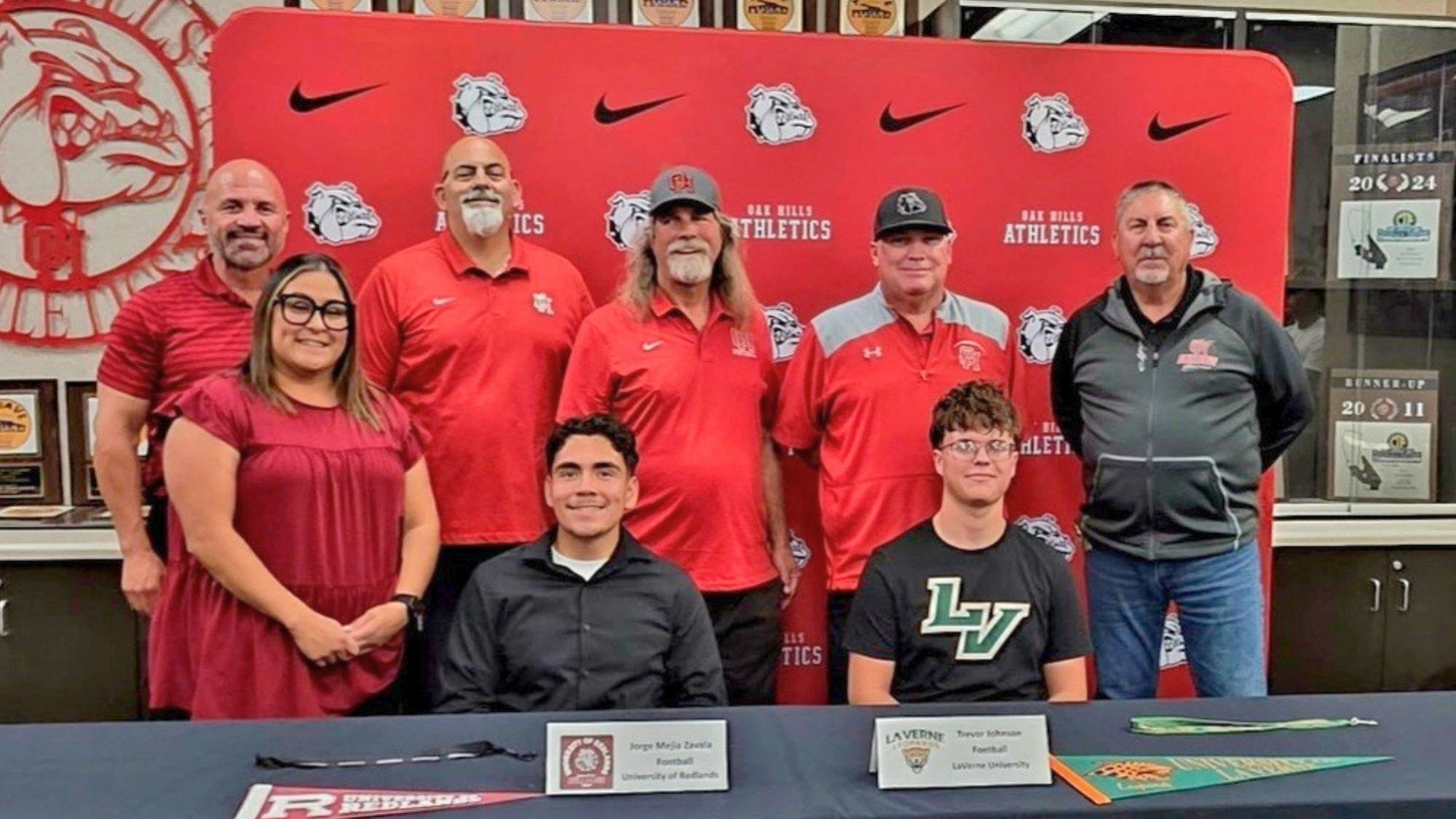 OakHillsAthletes Group of coaches, family members, and two student-athletes pose in front of an Oak Hills Athletics backdrop during a college signing event, with the students seated at a table displaying their university commitments.