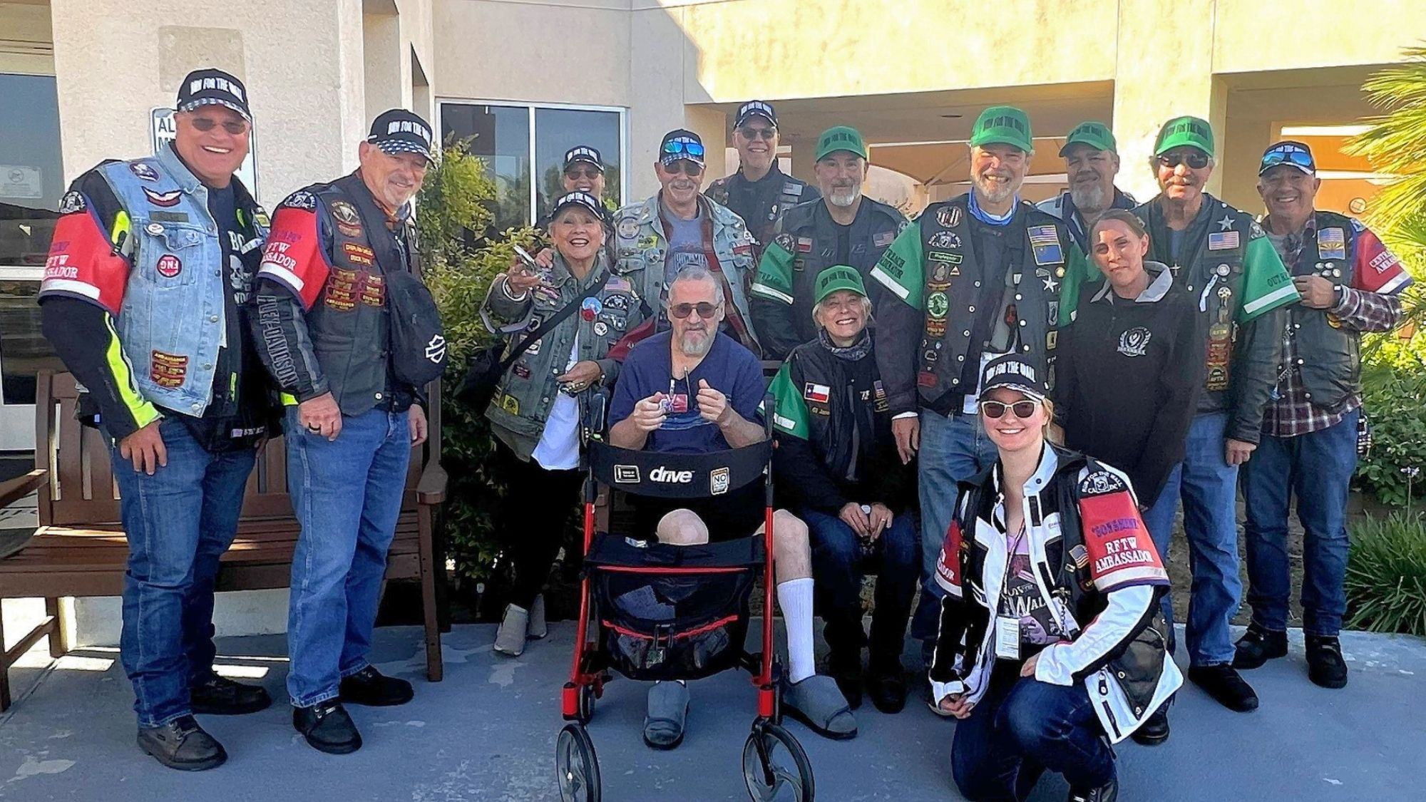 Group of motorcycle riders and veterans wearing patched riding vests and hats gathered outside a building.