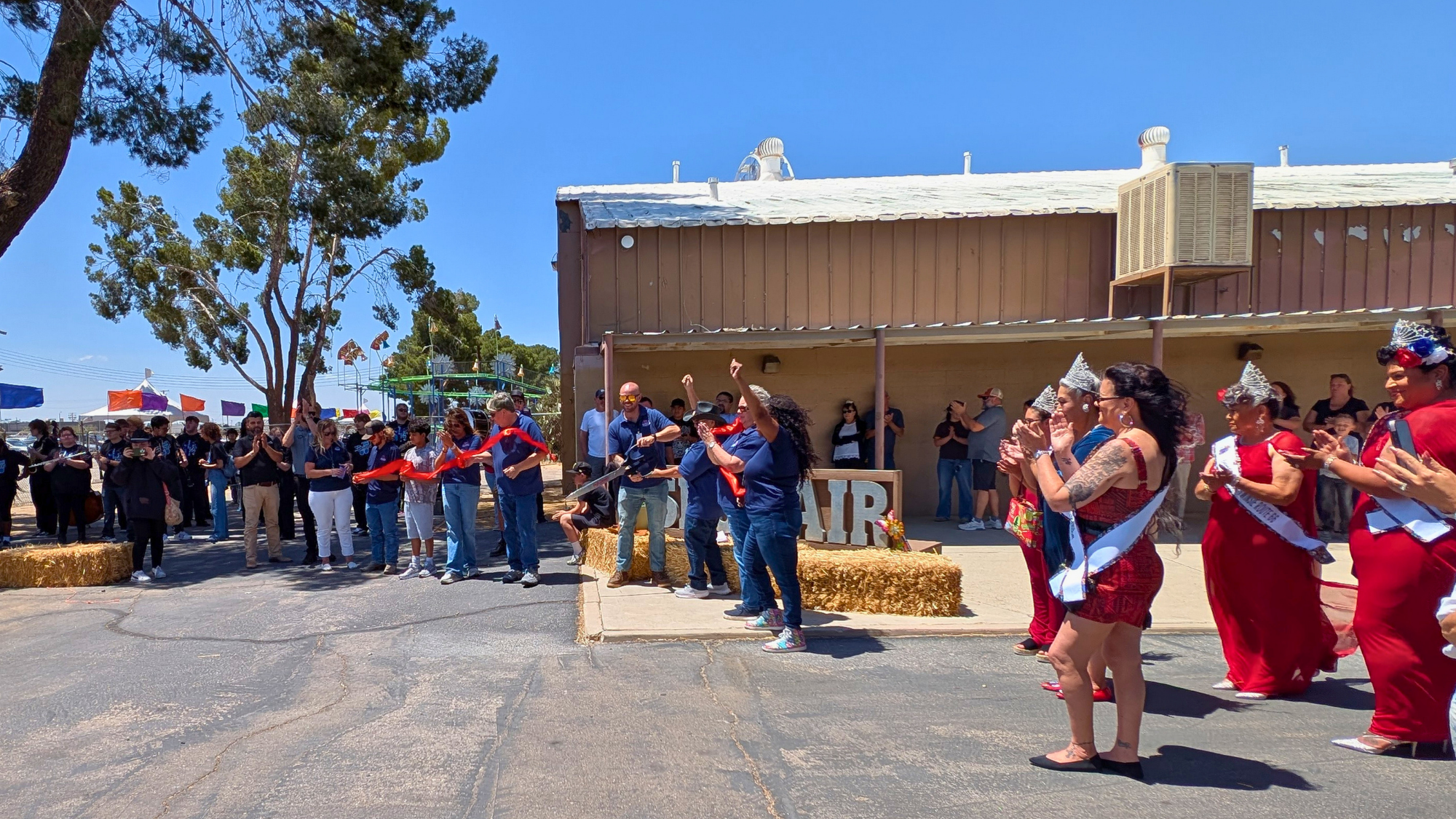 SBCFair A crowd gathers outside a fairgrounds building during a ribbon-cutting ceremony, with community members applauding as participants cut a red ribbon under a bright blue sky.