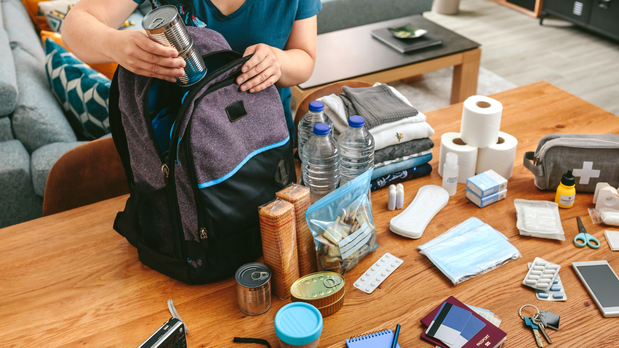 Person packing an emergency preparedness backpack with canned food and essential supplies on a table.