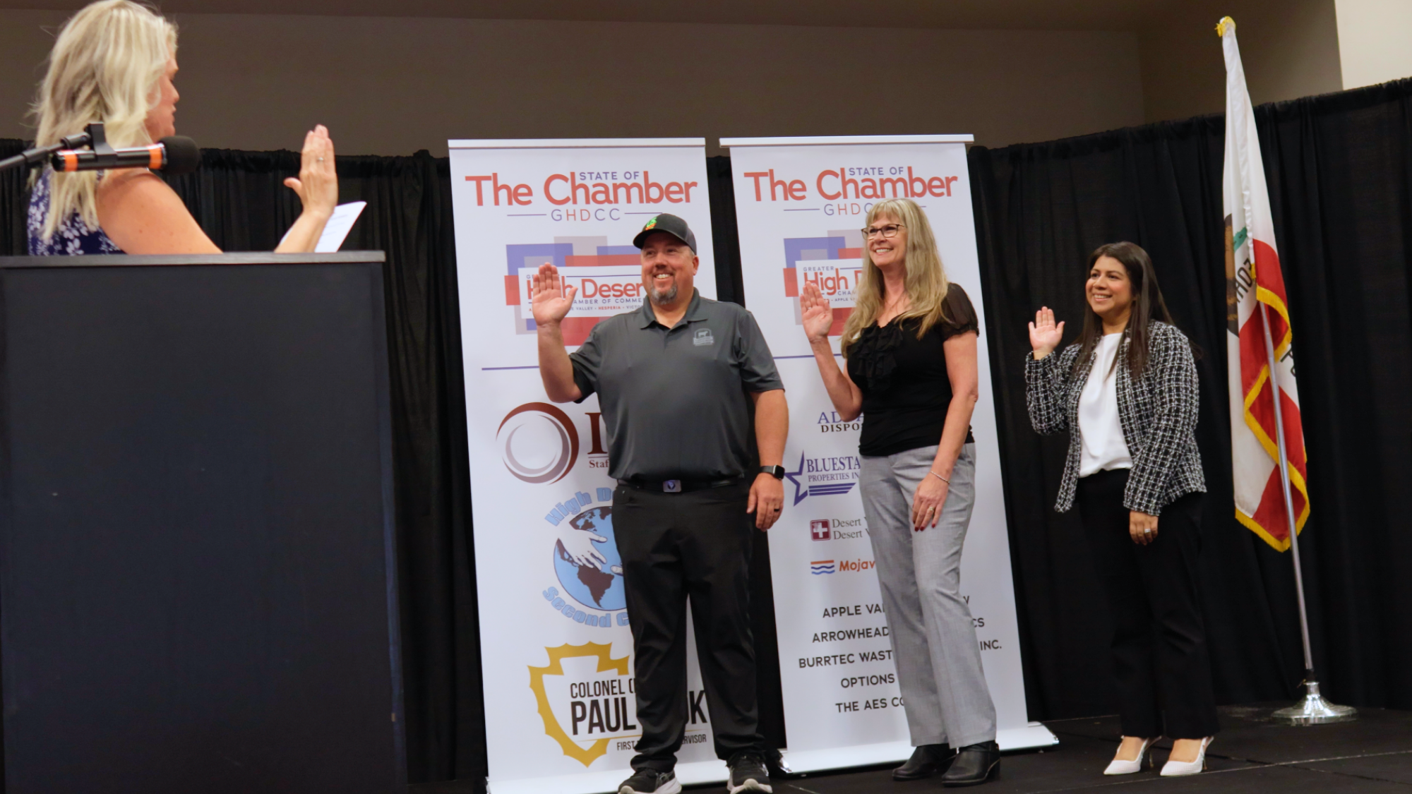 Three individuals stand on a stage with their right hands raised as they take an oath during a Greater High Desert Chamber of Commerce event, while a speaker administers the ceremony from a podium. Chamber banners and a California state flag are displayed in the background.