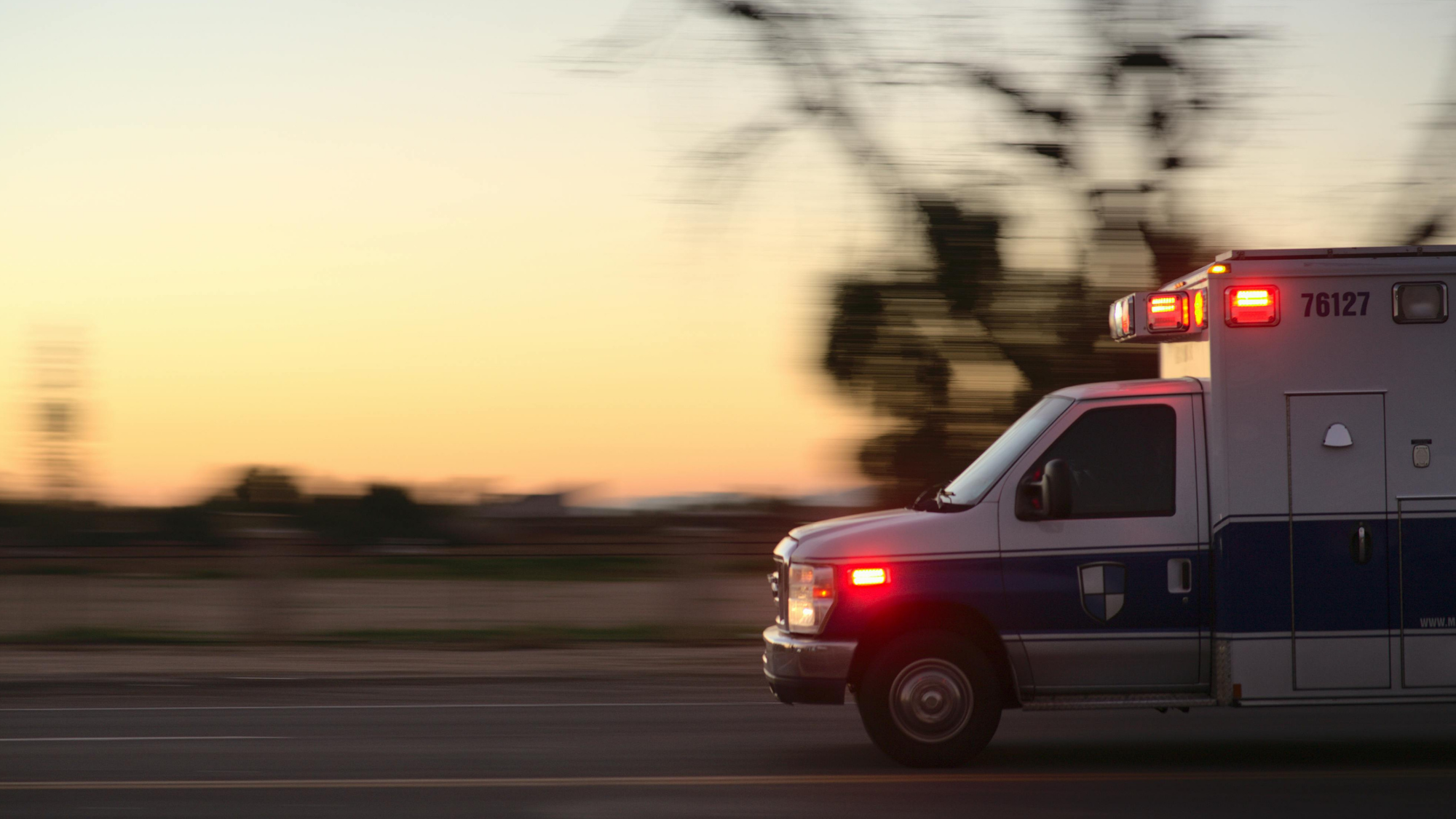 An ambulance with flashing red emergency lights speeds down a roadway at sunset. The vehicle is captured in motion, with the background blurred to emphasize its rapid response.