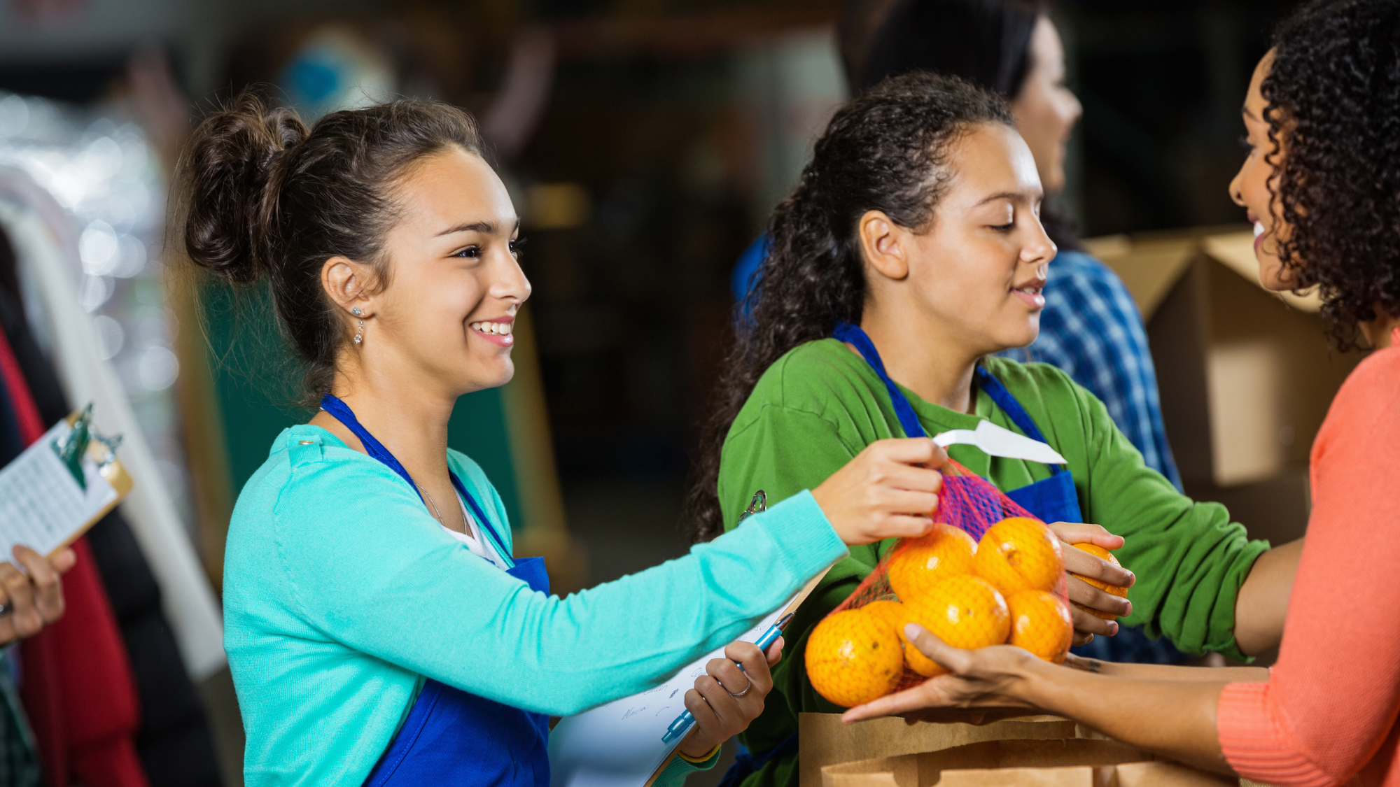 Volunteers wearing blue aprons smile as they distribute a bag of fresh oranges to a community member during a food distribution event. One volunteer holds a clipboard while another assists with groceries.