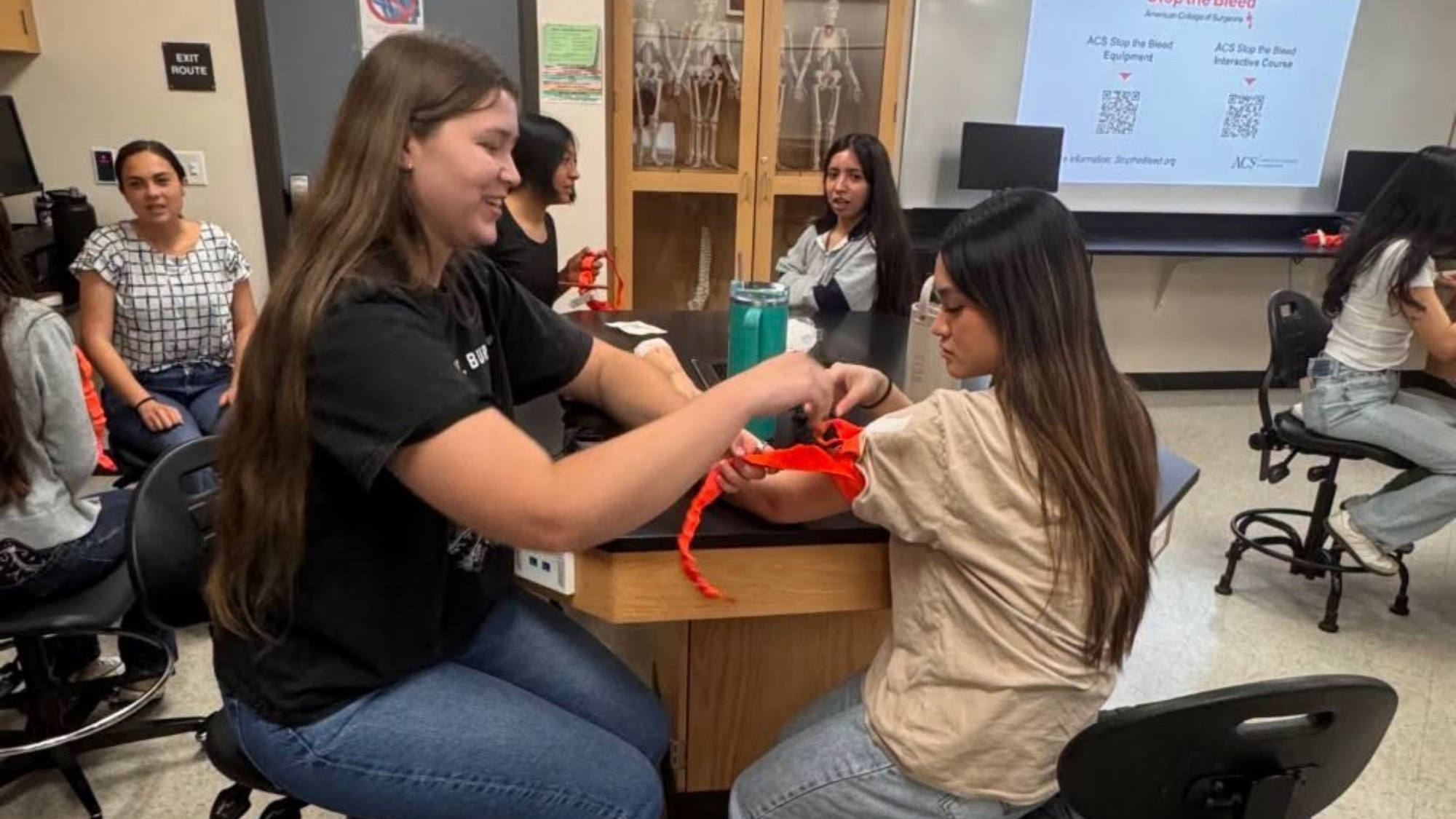 Students participate in a hands-on emergency preparedness training session in a science classroom. One student practices applying a tourniquet to another student’s arm while classmates observe and work in small groups.