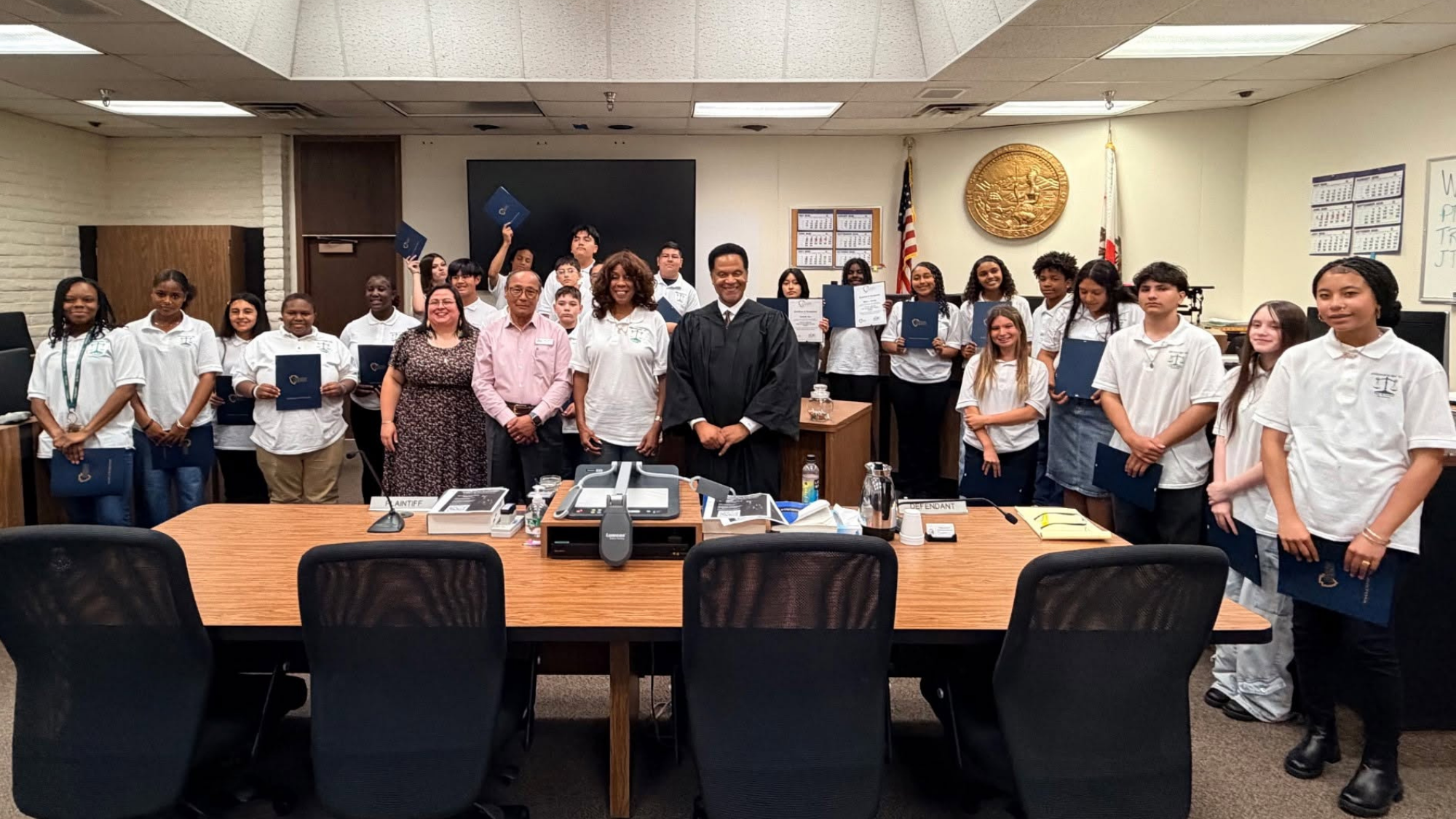 VictorvilleMockTrial Group photo inside a courtroom featuring students, educators, and legal professionals participating in a mock trial program. Several students hold certificates or folders while standing around the judge’s bench and conference table.