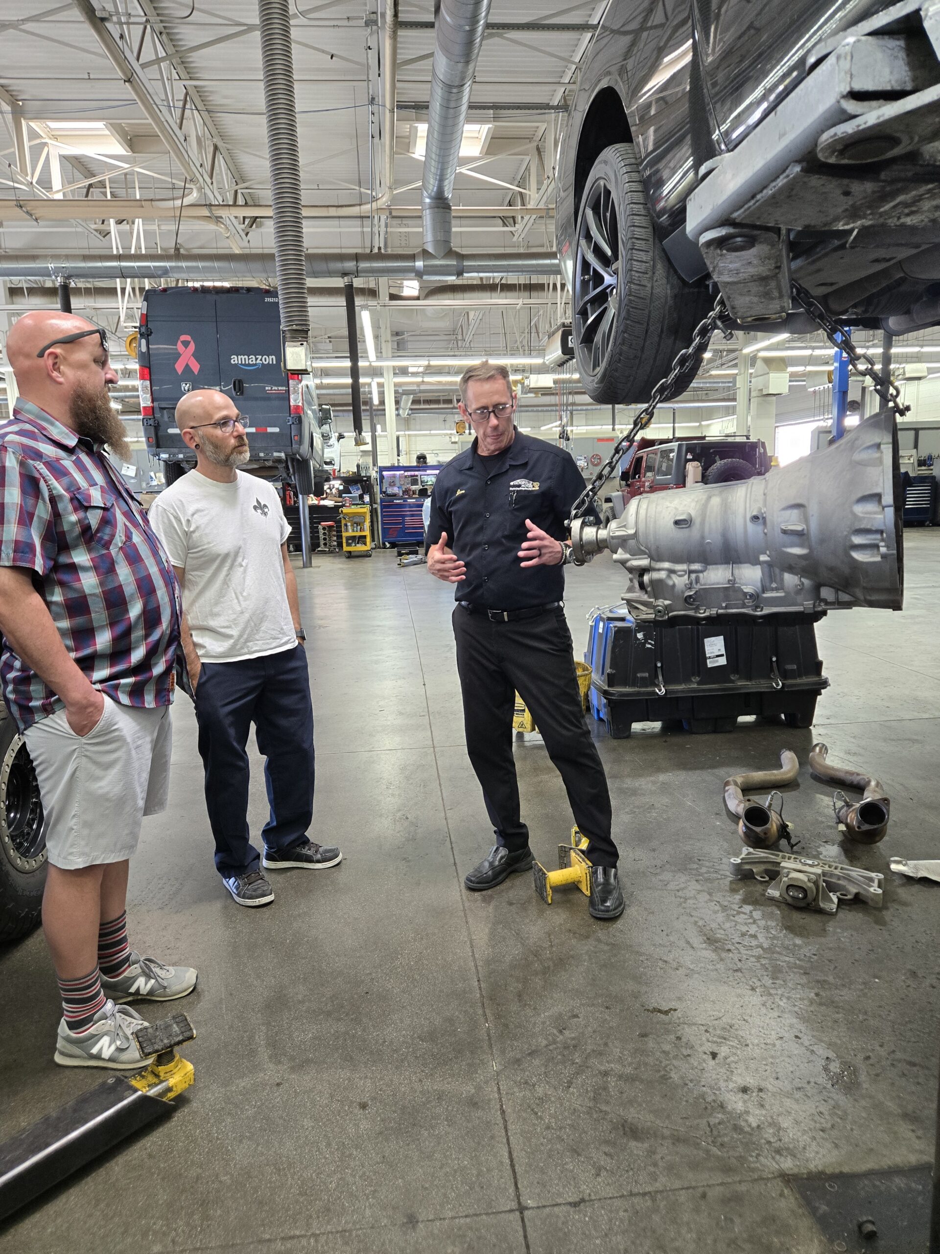 An automotive instructor explains drivetrain components to two visitors inside a vehicle maintenance shop.