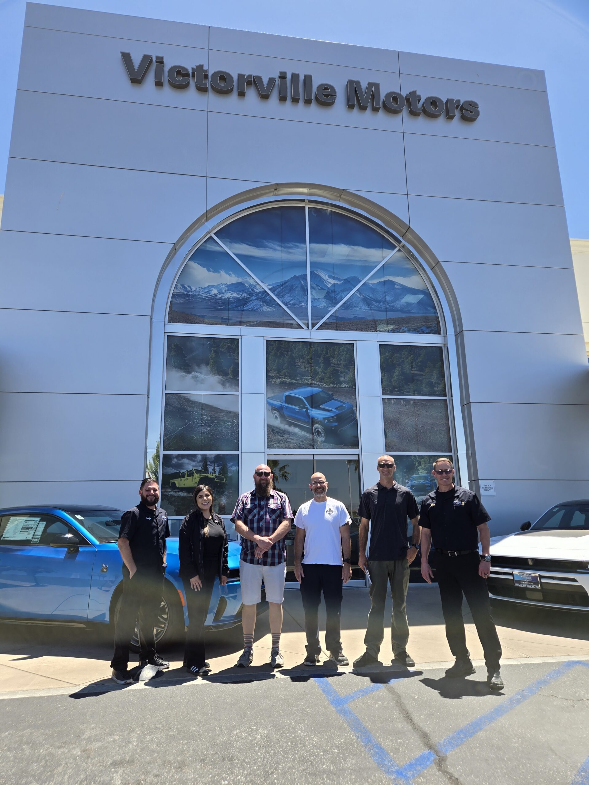 Six people stand outside the Victoryville Motors dealership in front of the main entrance for a group photo.