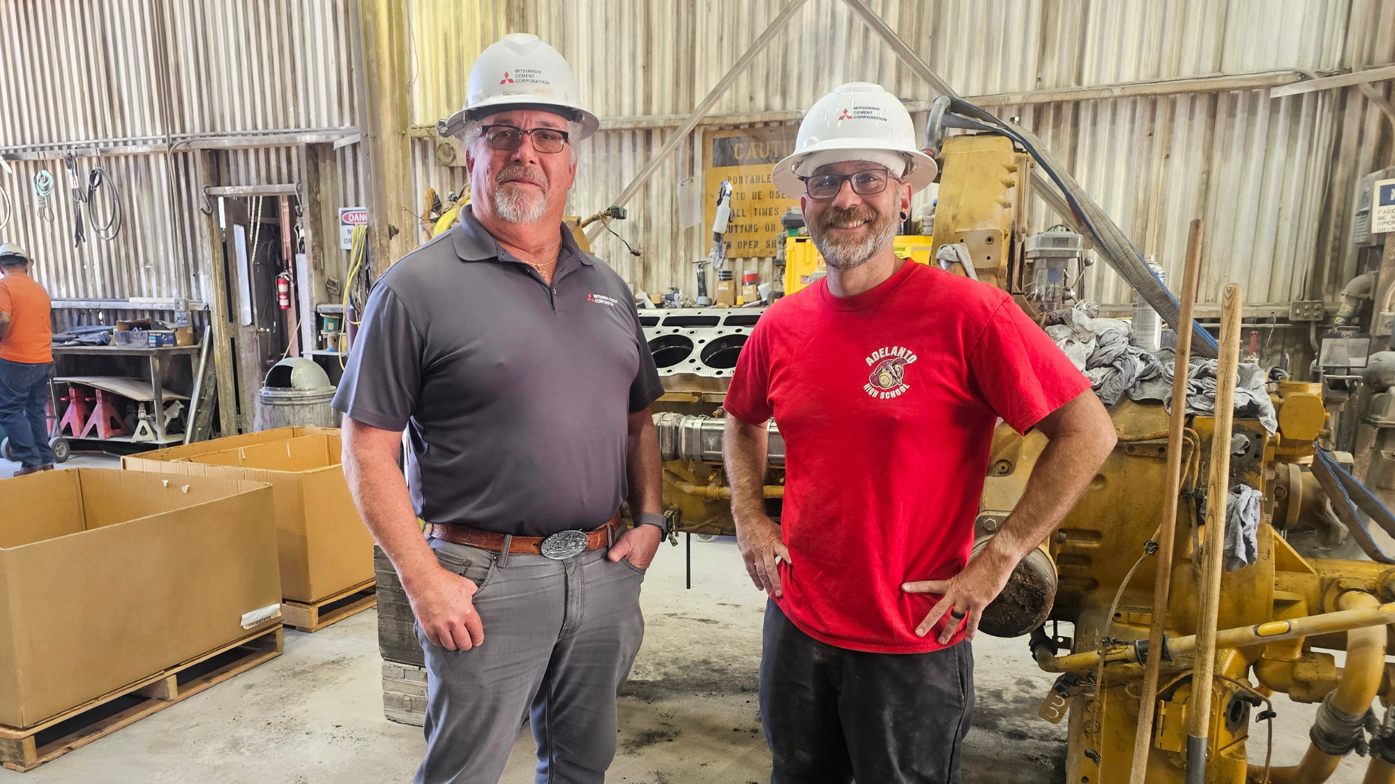 CTEExternship Two employees wearing white hard hats stand inside an industrial maintenance shop beside a large piece of heavy equipment.