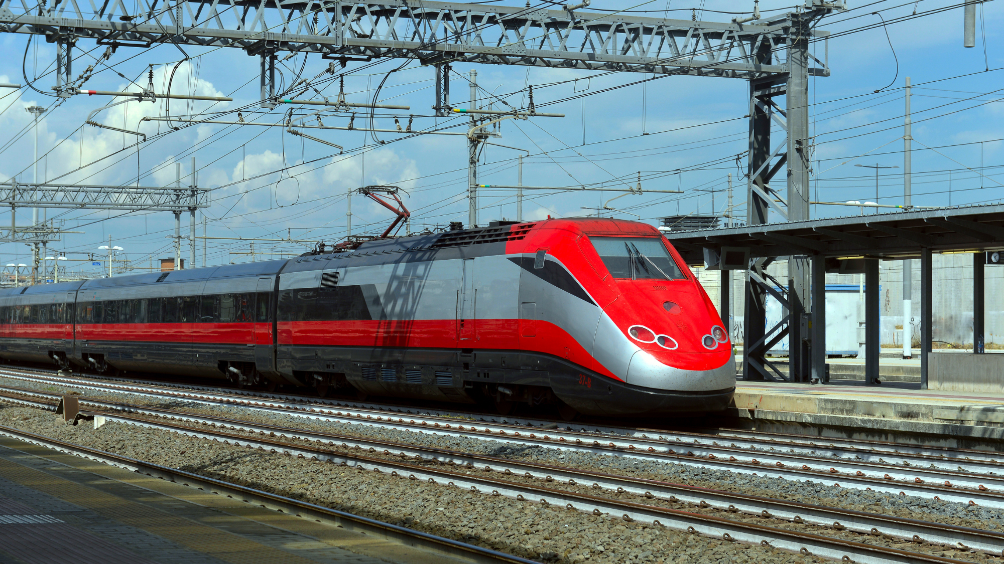 HighSpeedRail A modern red-and-silver high-speed passenger train sits at a railway station beneath overhead electric power lines.