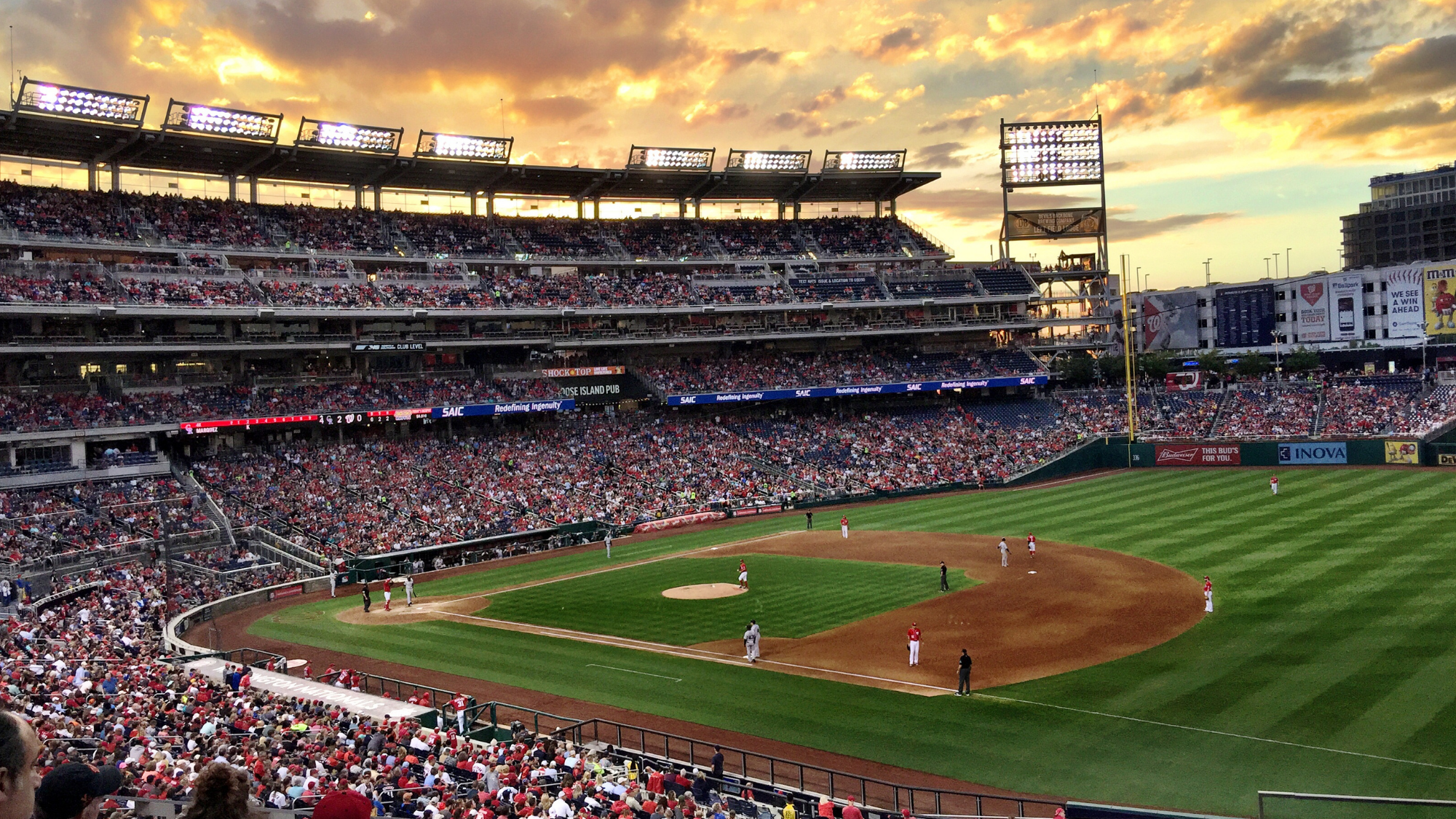 MLB2026 Wide view of a packed baseball stadium during a game at sunset, with players positioned on the field beneath glowing stadium lights.