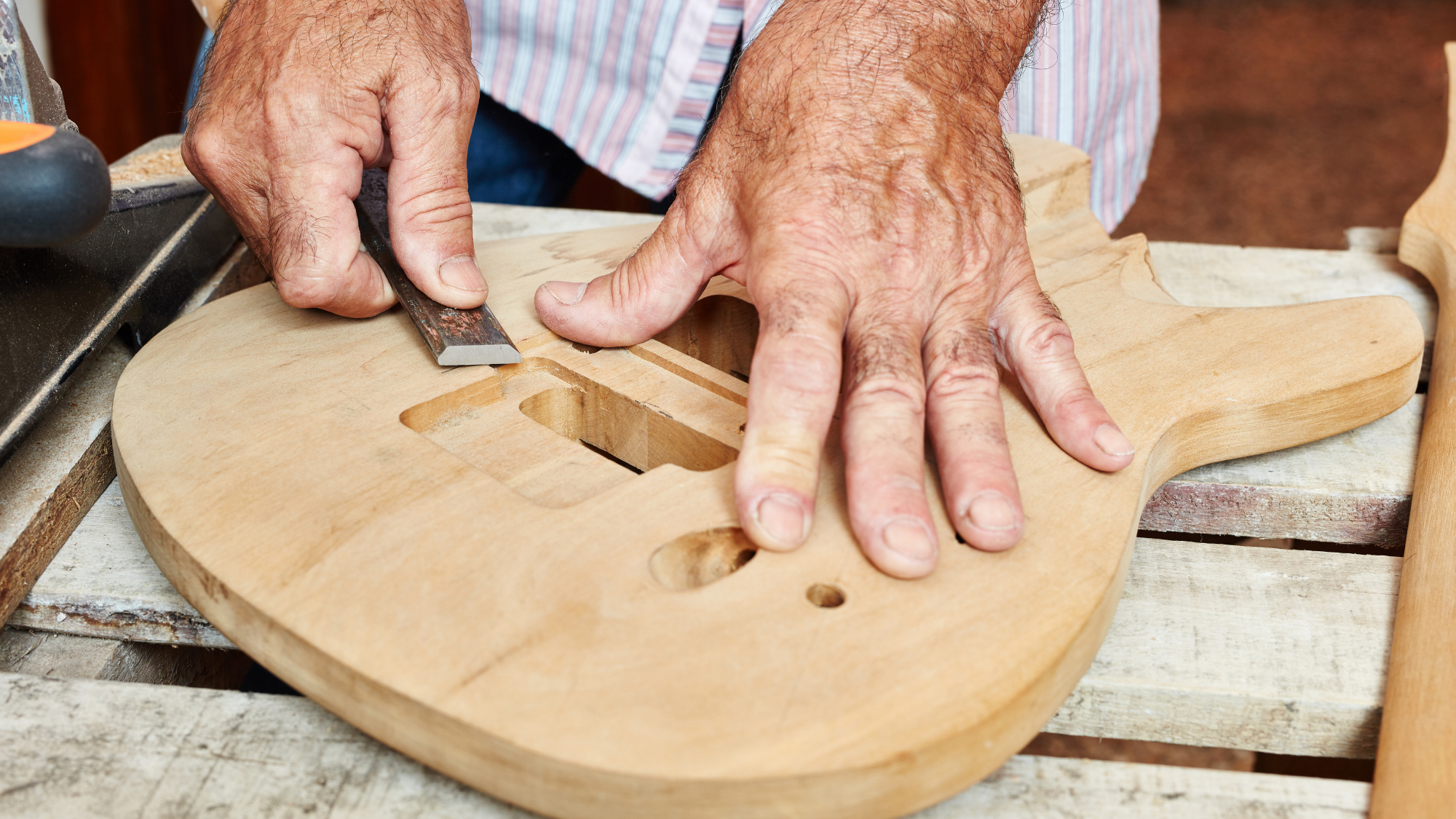 PatriotMusic Close-up of a craftsperson using a wood chisel to shape the body of an unfinished electric guitar.