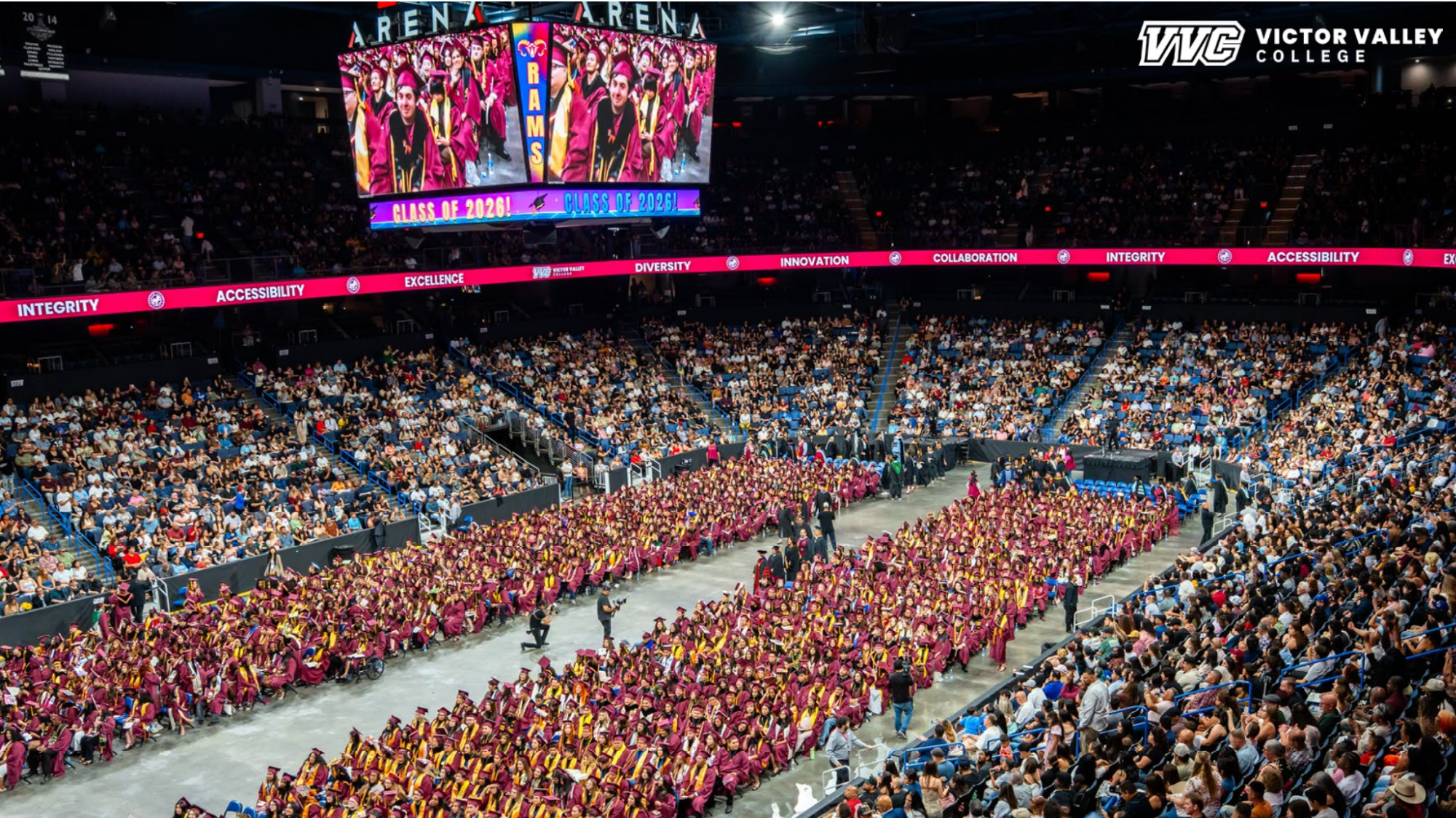 Wide view of Victor Valley College's 2026 commencement ceremony inside a packed arena. Hundreds of graduates wearing maroon caps and gowns are seated on the arena floor while family, friends, and supporters fill the surrounding stands.
