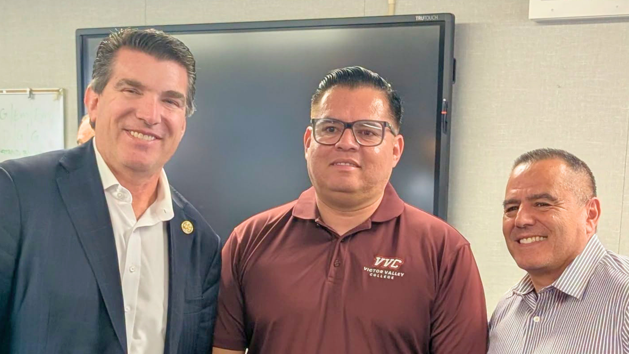 Three professionals stand together smiling during a meeting at Victor Valley College. One person wears a Victor Valley College polo shirt, while the others are dressed in business attire.